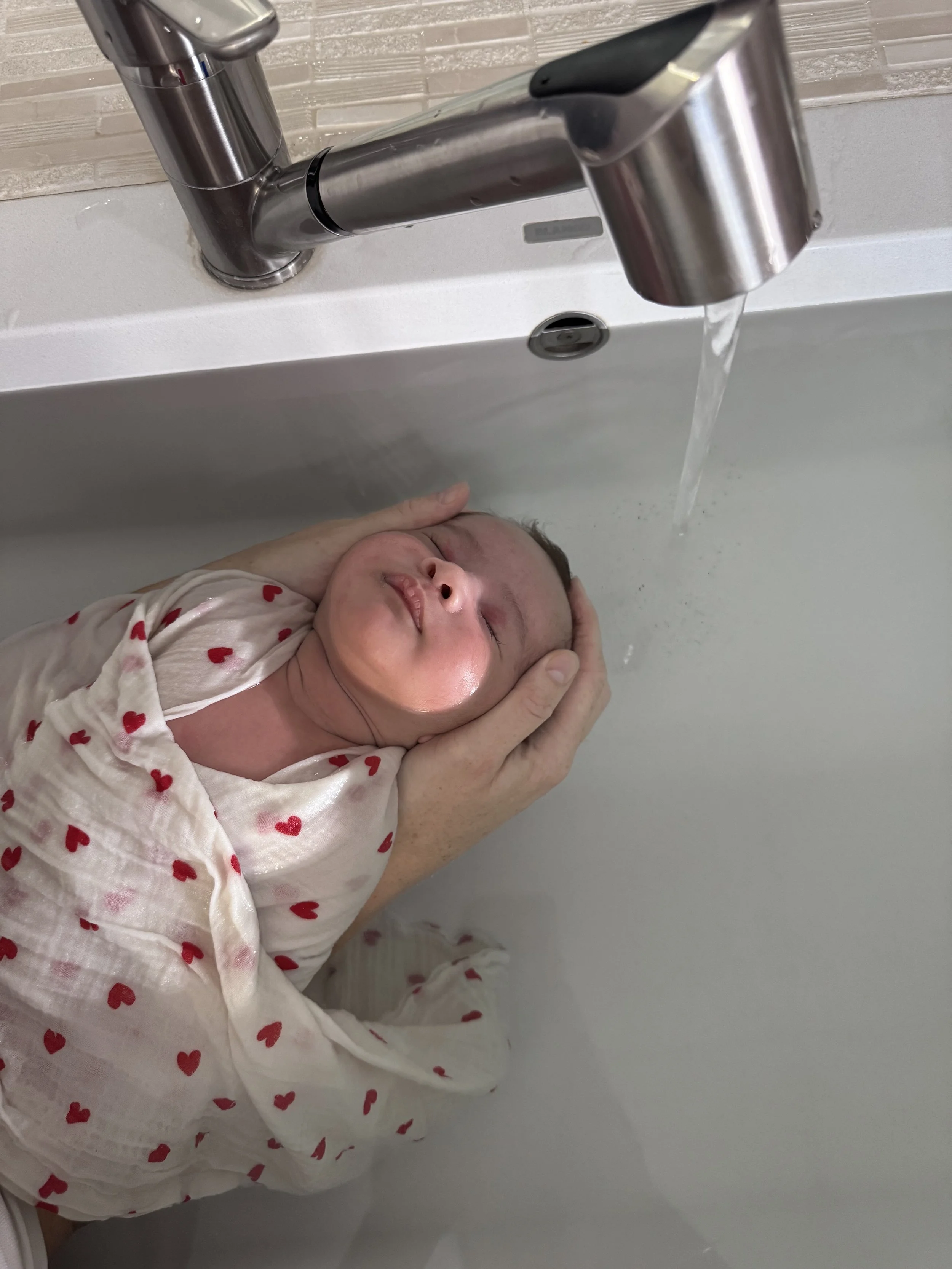Un bébé avec des vêtements à motifs de petits cœurs rouges, la tête appuyée contre la main d’un adulte, sous un robinet d’évier avec de l’eau qui coule.