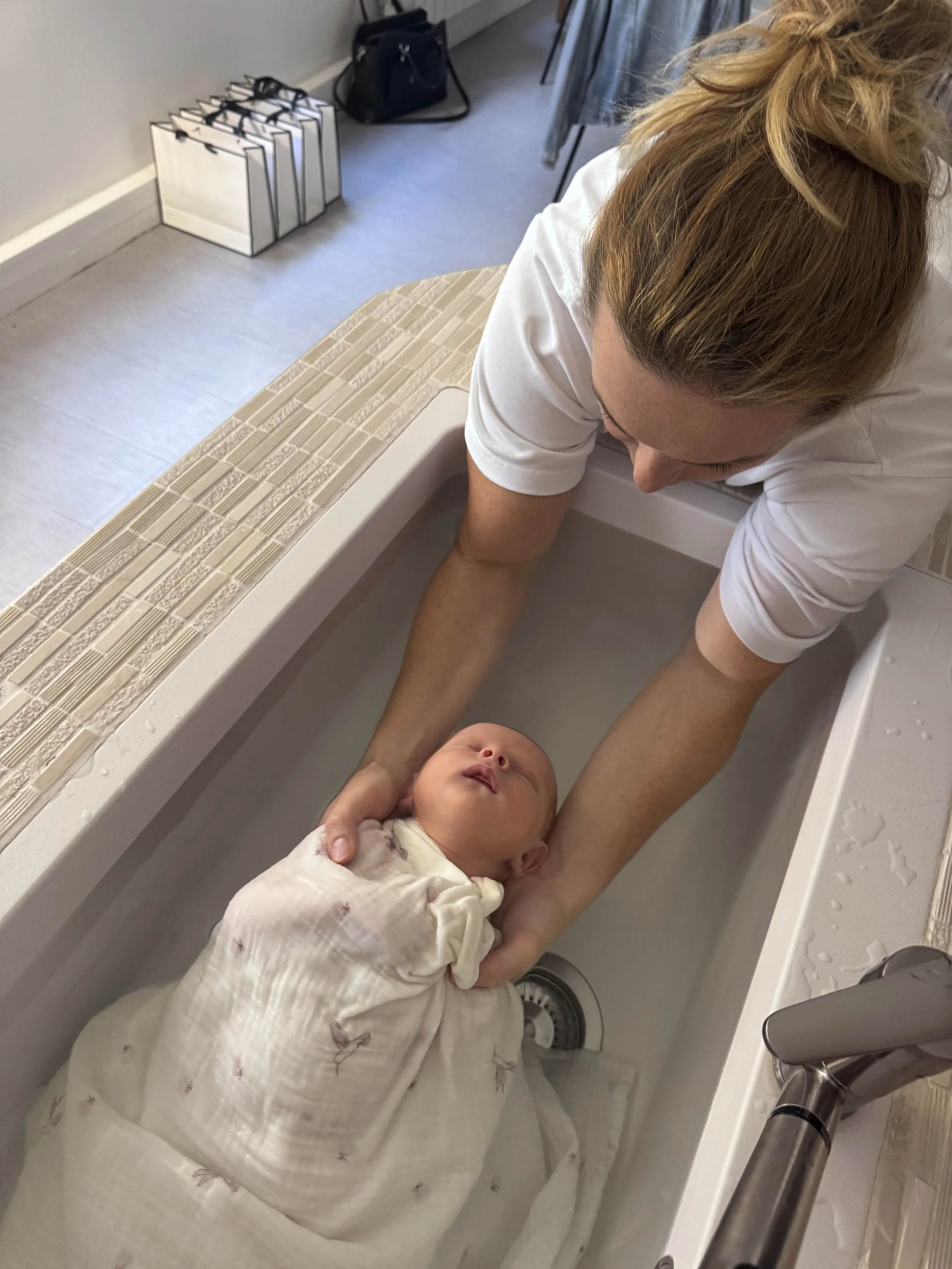 Une femme donne un bain à un bébé dans une baignoire. La femme porte un t-shirt blanc et le bébé est enveloppé dans une couverture blanche avec des motifs roses.
