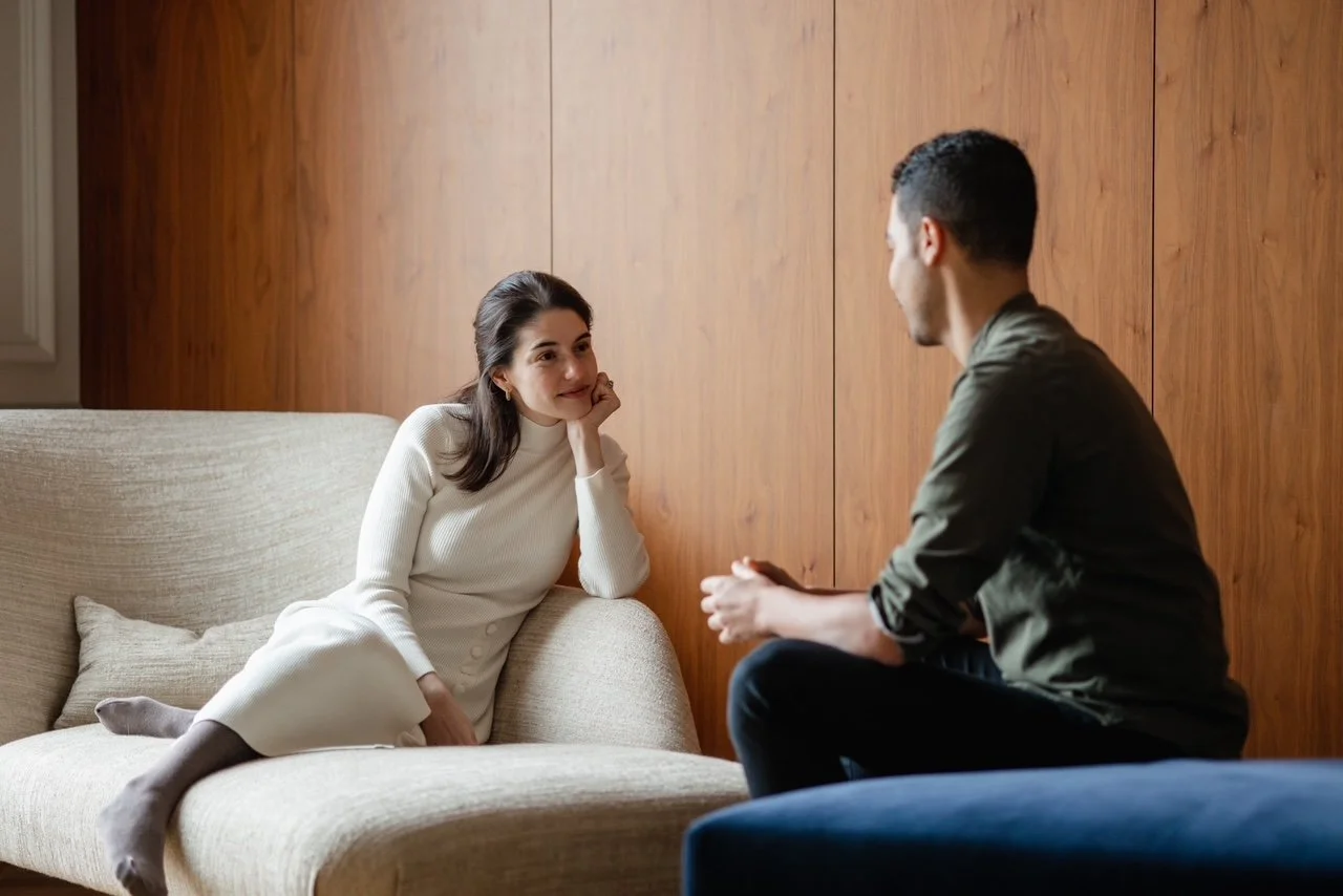 Une femme assise sur un canapé beige, écoutant un homme qui parle, dans une pièce aux murs en bois.