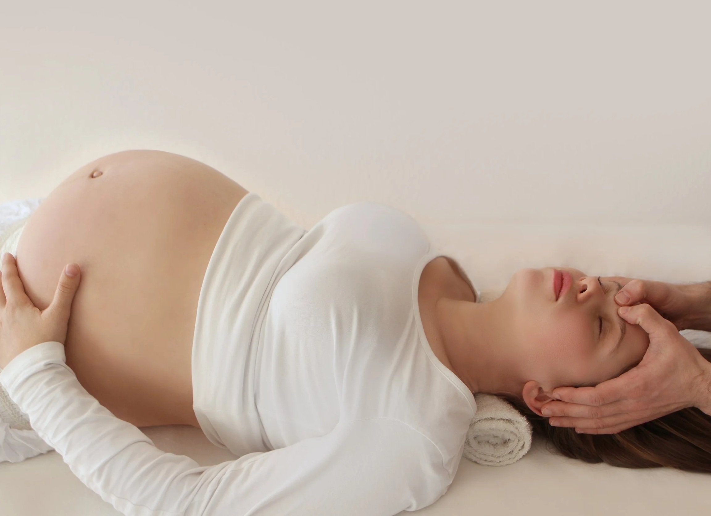Une femme allongée sur le dos, semble être enceinte, portant un t-shirt blanc. Elle se repose les yeux fermés, tenant son ventre avec une main, dans un cadre paisible.