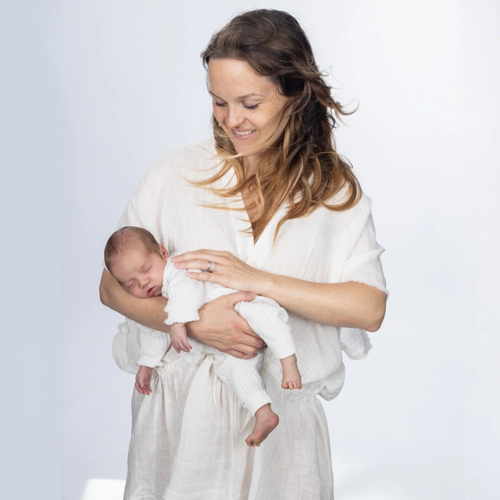 Une femme souriante tient un bébé endormi dans ses bras, tous deux habillés en vêtements blancs, contre un fond blanc.