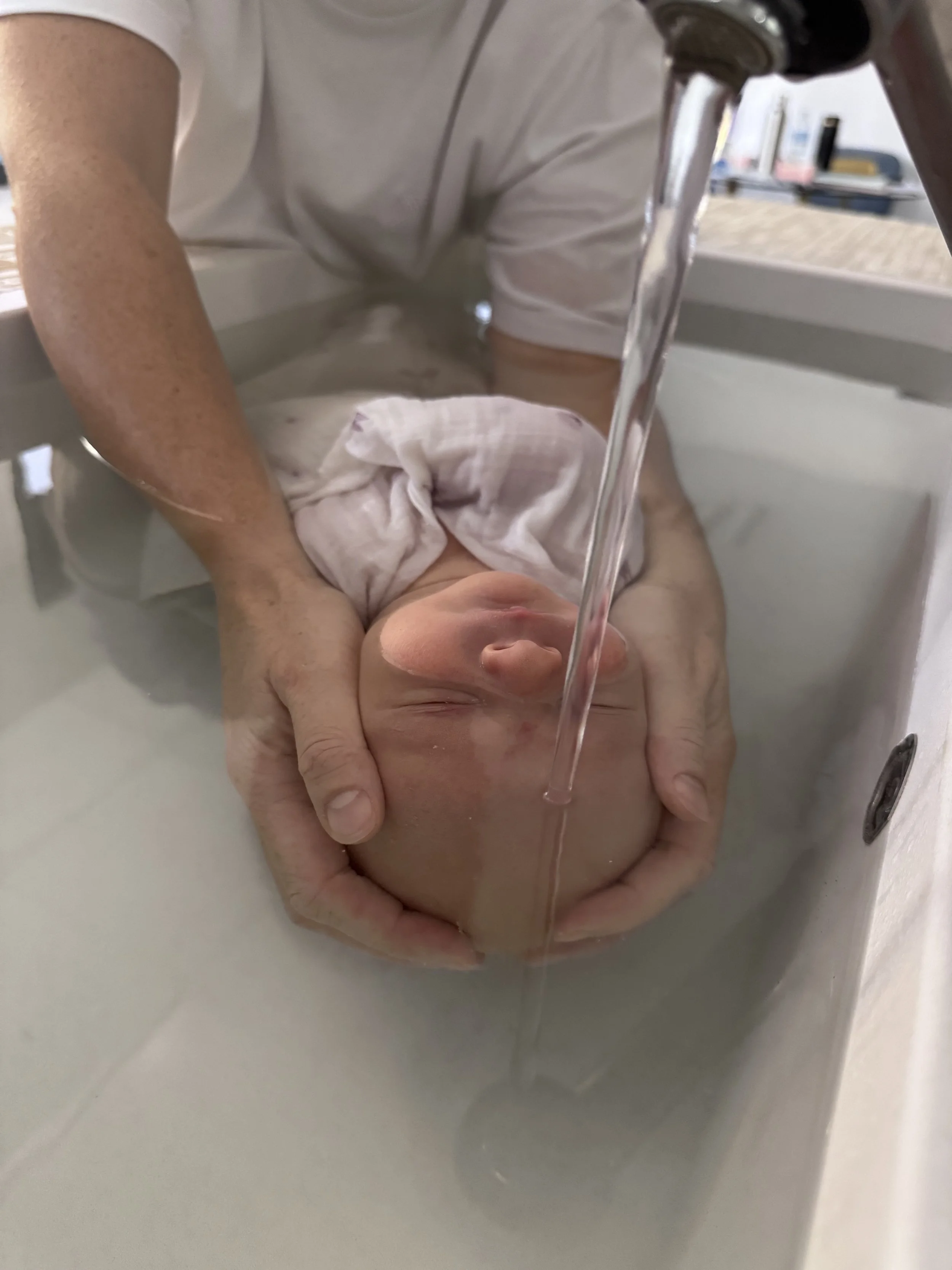 Une personne tient un bébé dans une bassine, le bébé est recouvert partiellement de tissu, et de l'eau coule du robinet.