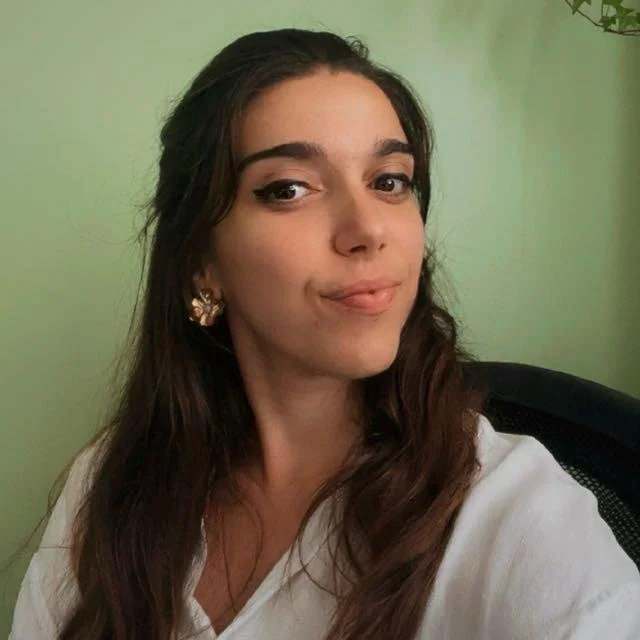 Justine Drochon à la Bulle Périnatale de Puteaux Jeune femme aux cheveux bruns, portant un chemisier blanc et des boucles d'oreilles en or, assise devant un mur vert pâle.