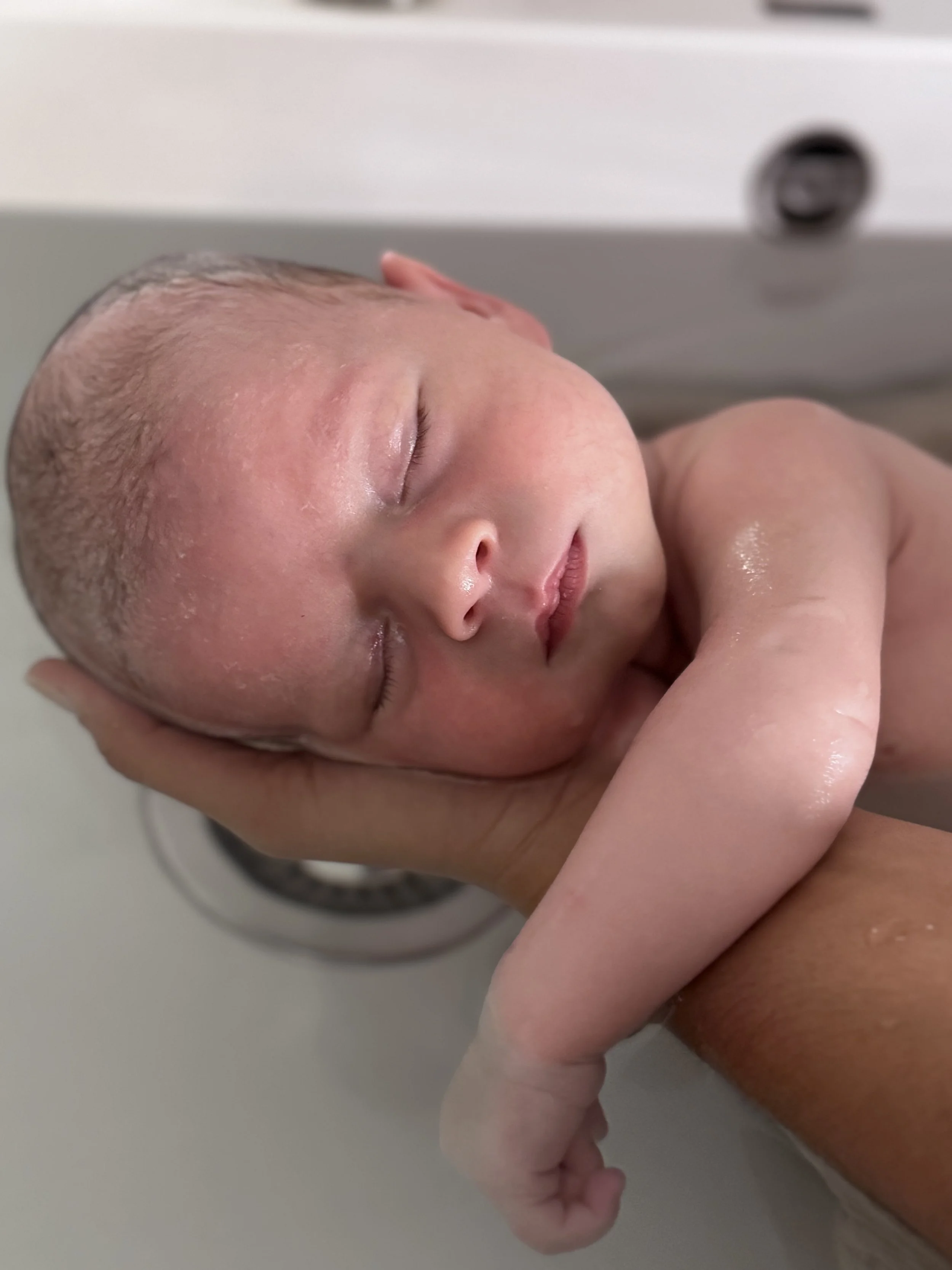 Un nouveau-né couché dans un bain, son visage en gros plan, yeux fermés, peau humide et douce.