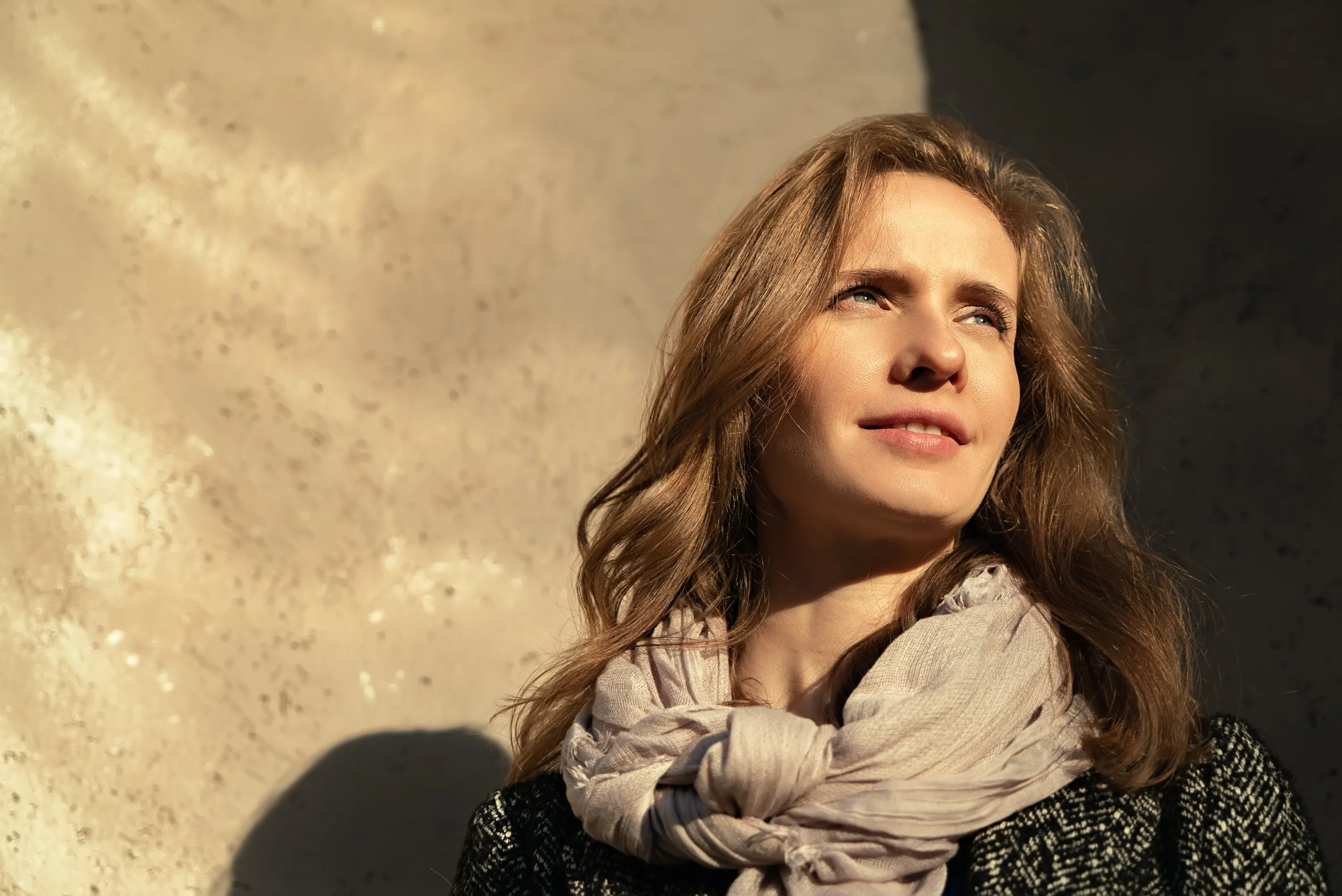 Une femme avec des cheveux longs et bruns regarde vers le haut, avec un fond clair et sombre en lumière naturelle.