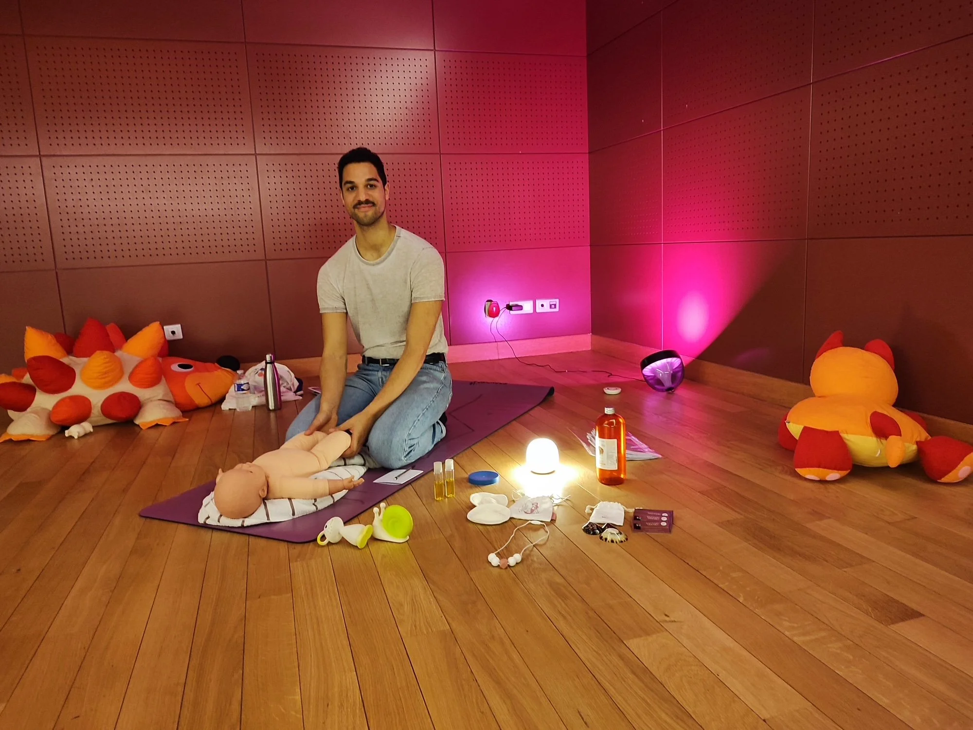 Un homme assis sur un tapis, entouré de jouets et d'accessoires pour bébés, dans une pièce au mur en bois rose avec des lampes colorées.