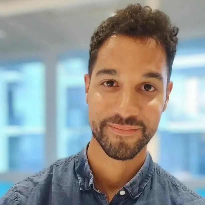 Matthieu Merabli à la Bulle Périnatale de Puteaux. Portrait d'un homme souriant, avec des cheveux bouclés, une barbe courte, portant une chemise en jean, en intérieur avec des fenêtres en arrière-plan.