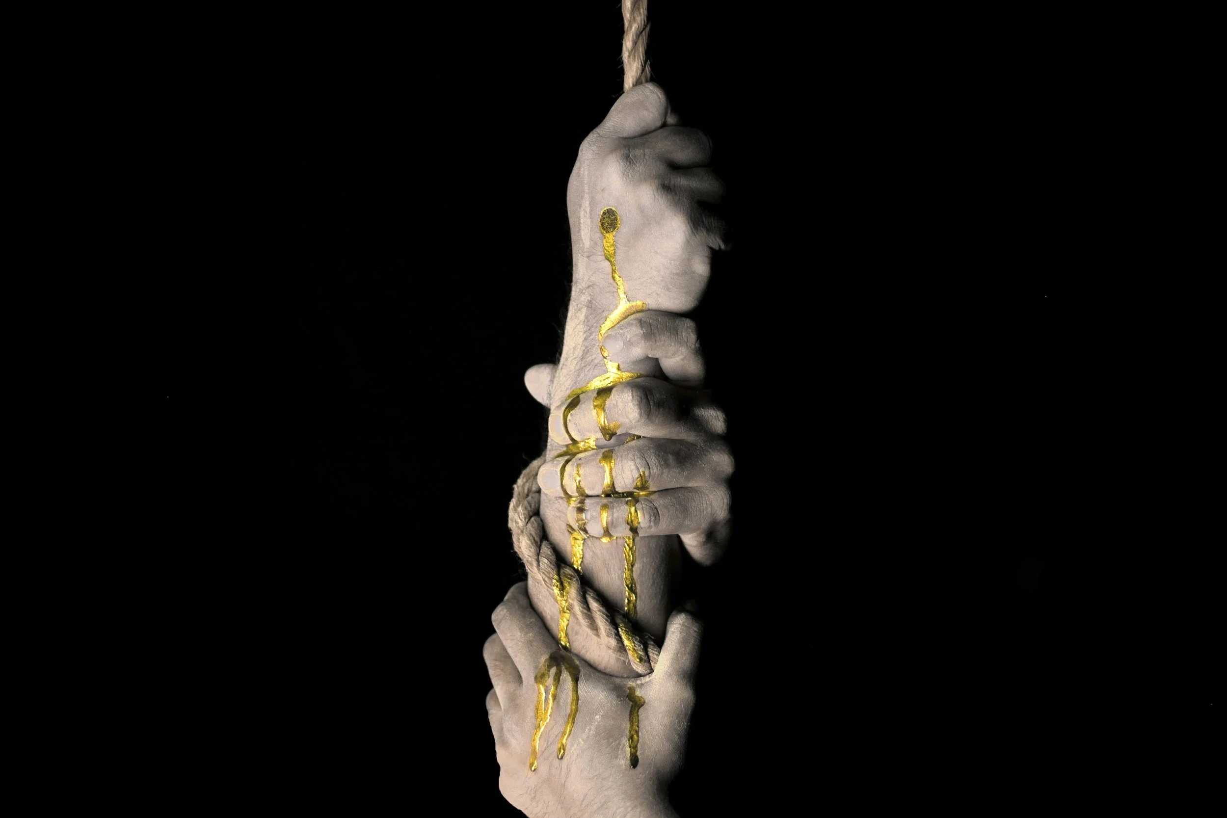 A white hand with visible veins and wrinkles holds a thick, twisted rope against a black background. Golden paint or gilt is dripped and splattered along the hand's fingers, knuckles, and wrist, creating a striking contrast.