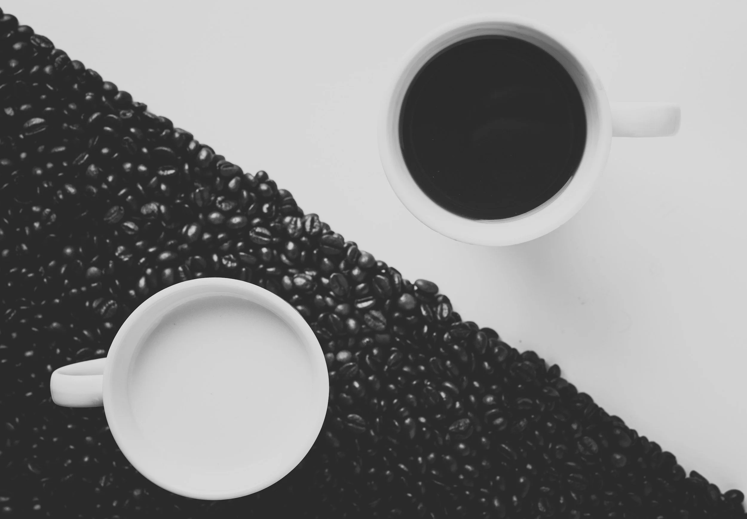 Black coffee beans scattered on a white surface with two white coffee cups, one filled with black coffee and the other empty.