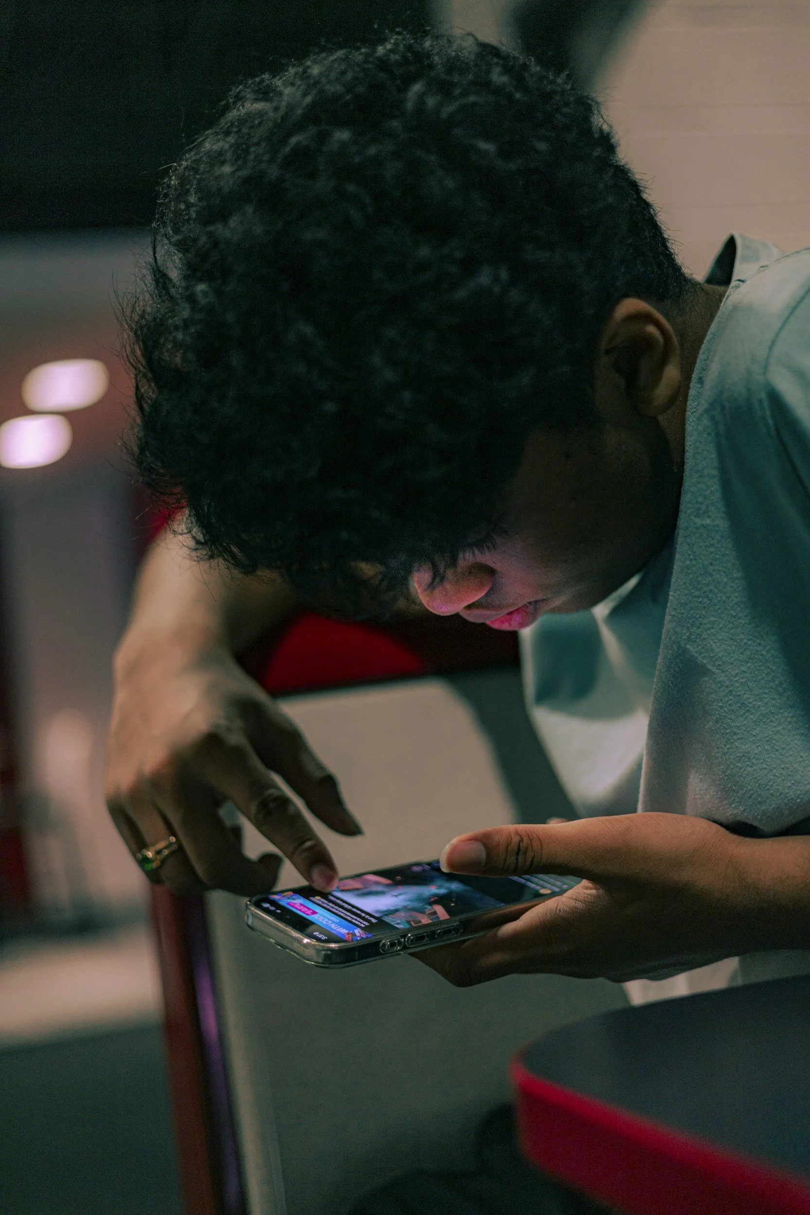 A young person with curly black hair, wearing a light-colored shirt, is leaning over a smartphone, tapping and scrolling on the screen, with a blurred background.