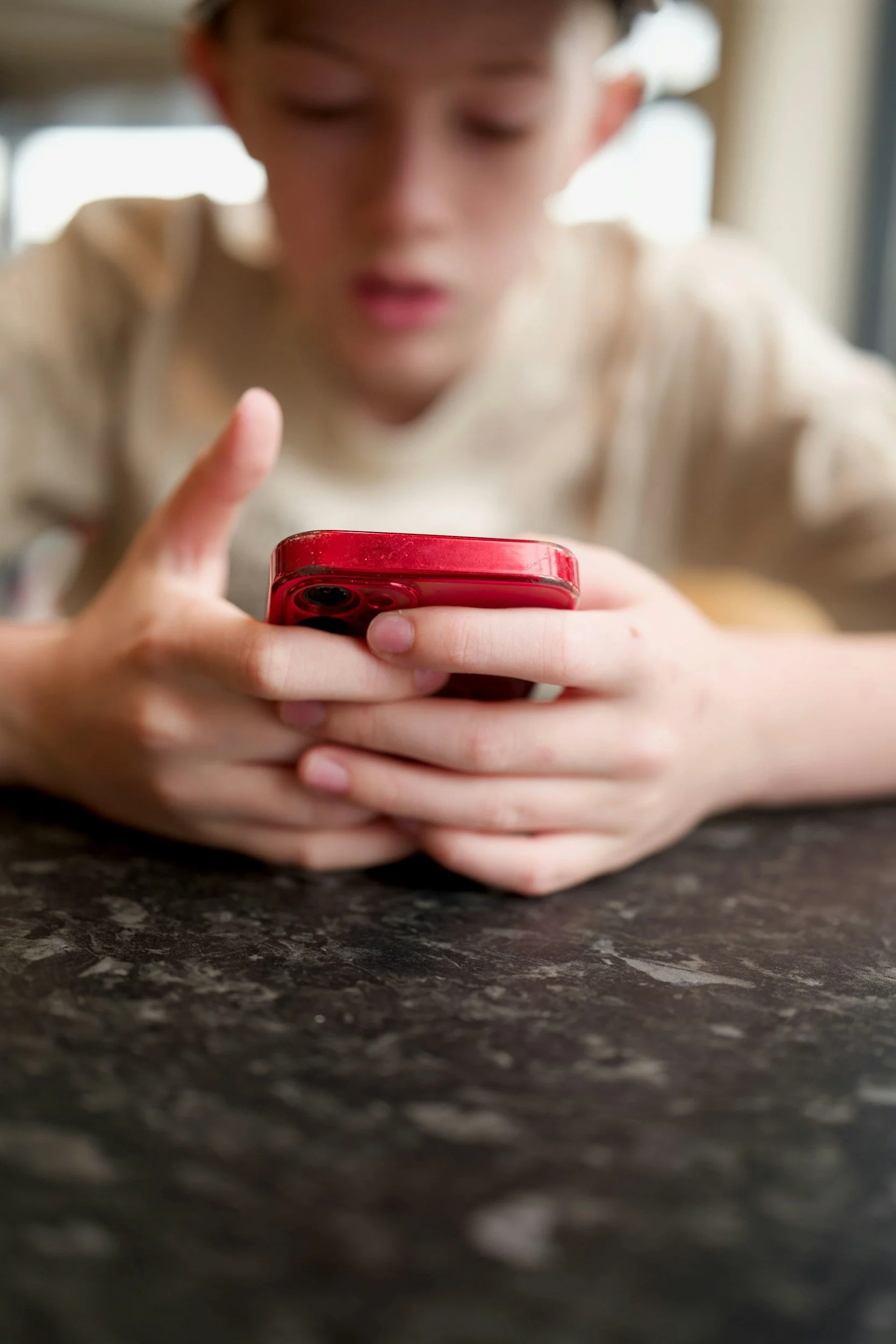 A young person with light brown hair, wearing a beige shirt, looking down at a red smartphone while sitting at a dark countertop.