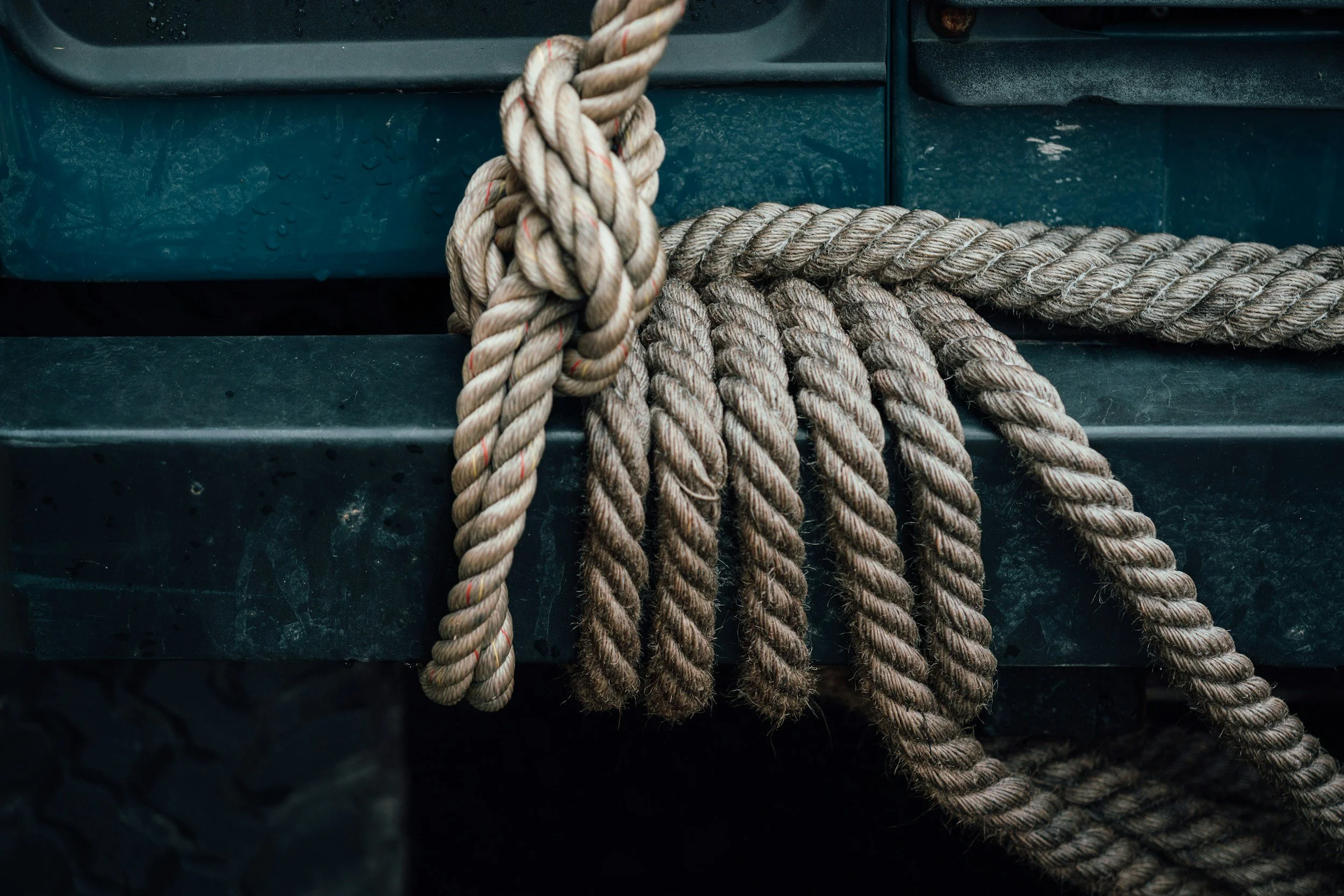 Close-up of beige and brown nautical ropes tied around a dark green surface, with water droplets on it.