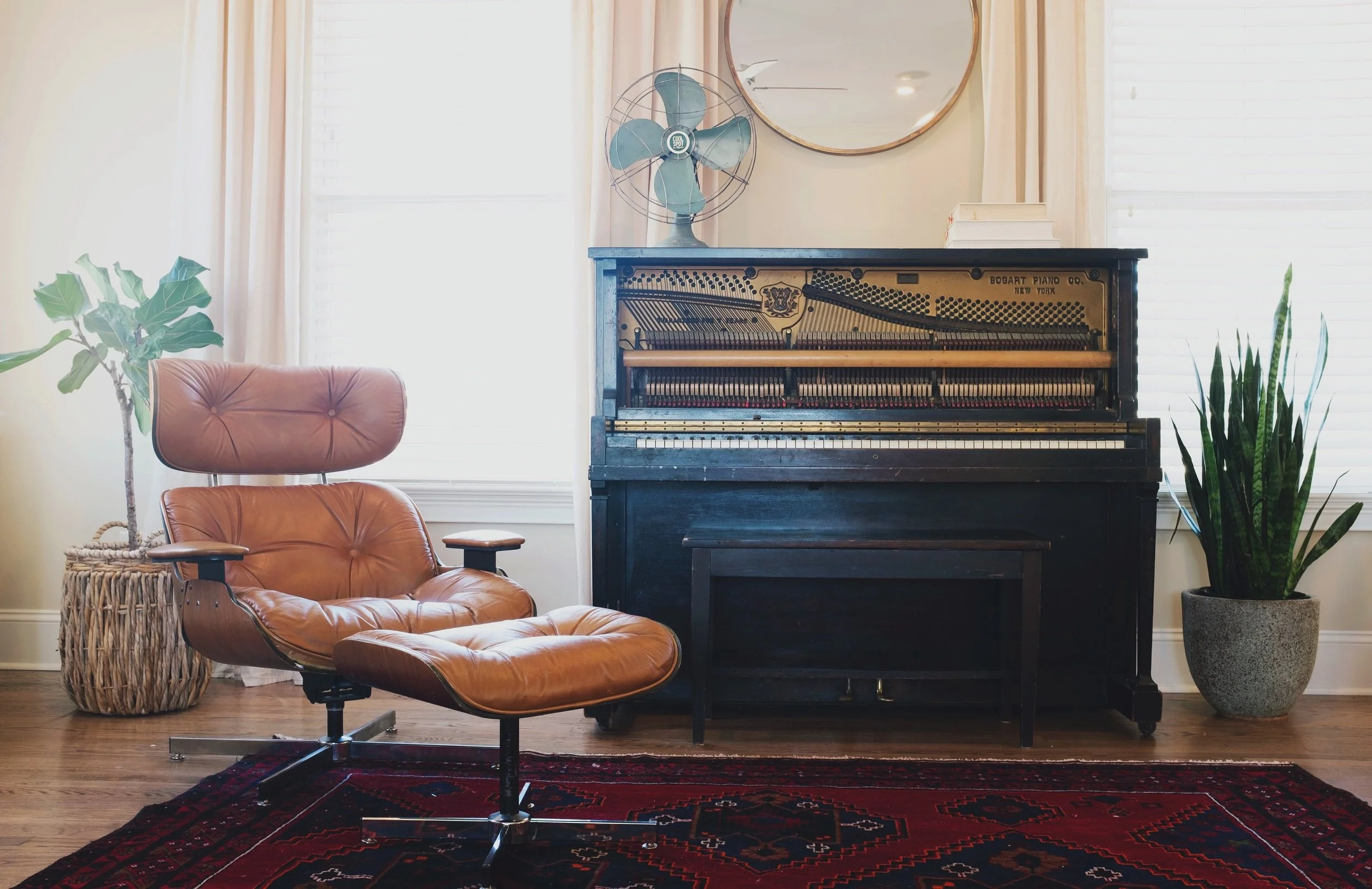 Ein Wohnzimmer mit einem schwarzen alten Klavier, einem braunen Sessel, einem grünen Pflanzenübertopf, einer Leuchterlampe, einem runden Spiegel und einem Vintage Tischventilator.