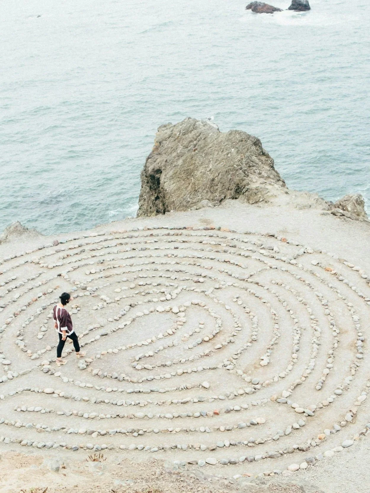 Eine Person beobachtet ein spiralförmiges Steingartenmuster am Meer, mit großen Felsen im Wasser im Hintergrund.