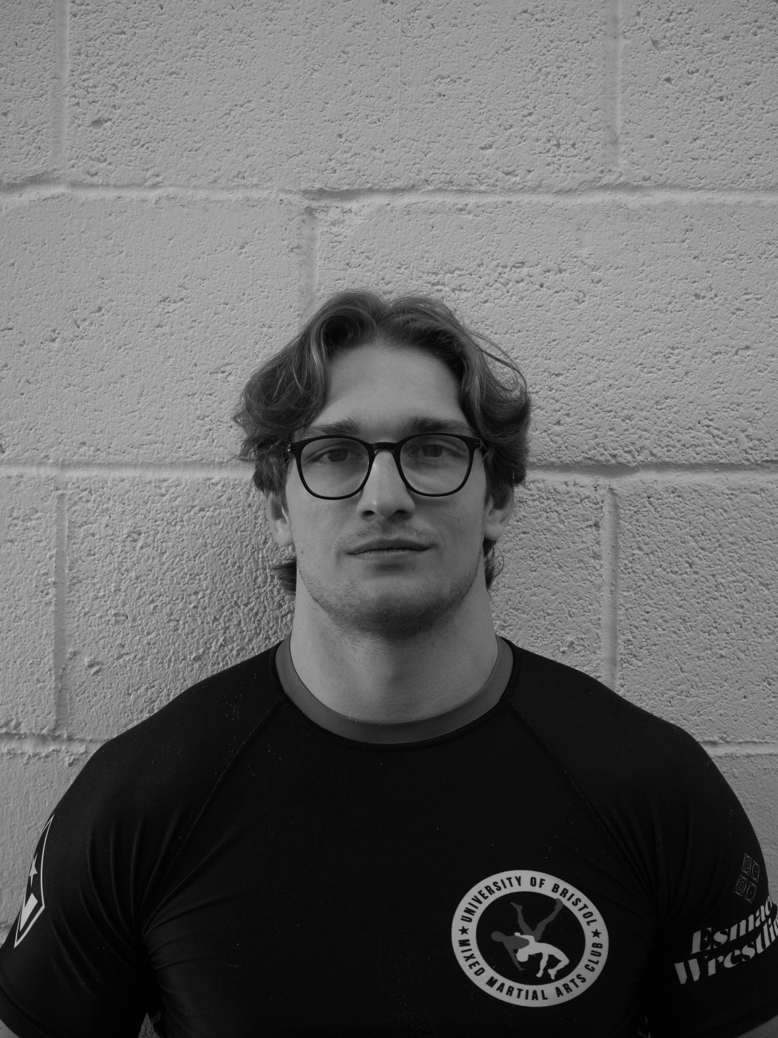 Black and white photo of a young man with glasses, curly hair, wearing a sports shirt with a martial arts club logo, standing in front of a textured brick wall.