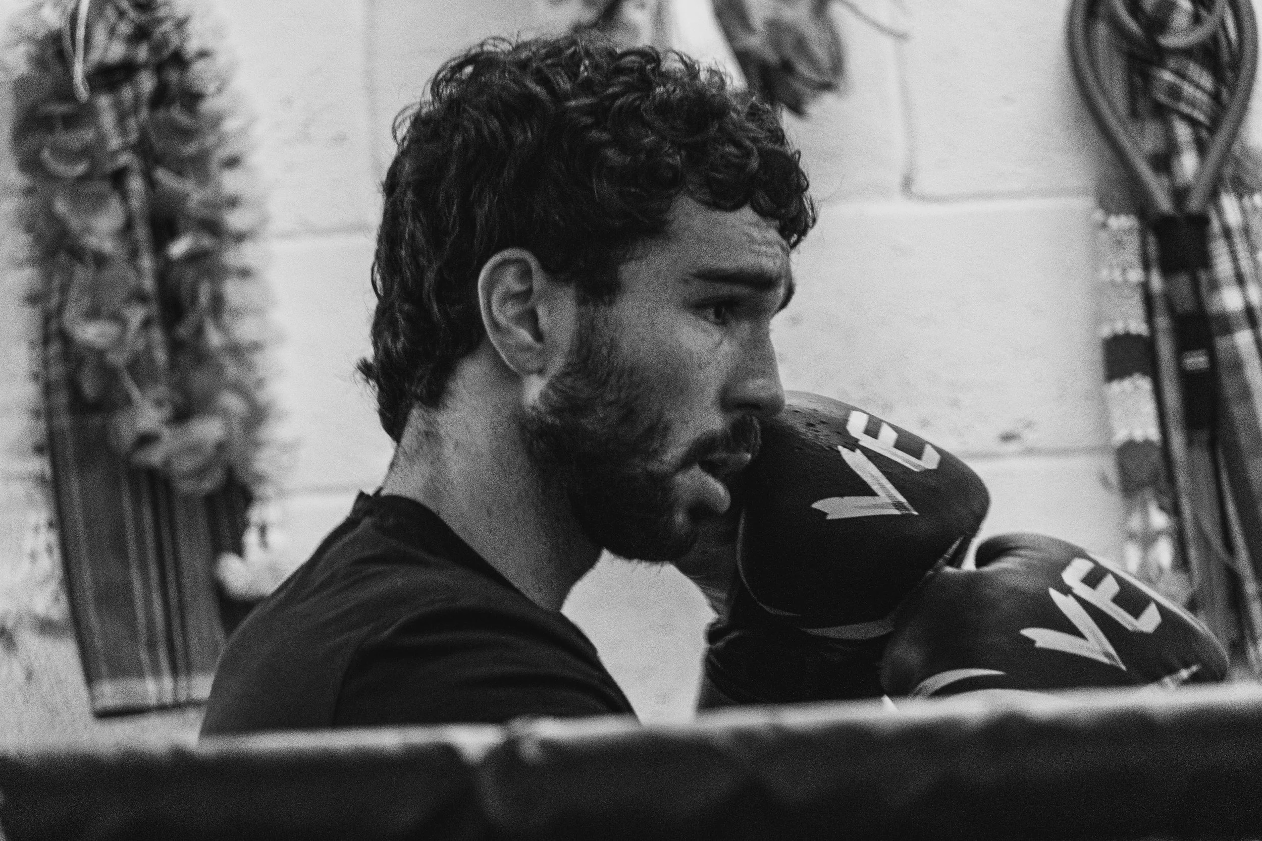 A black and white photo of a young man with curly hair and a beard, sitting with his eyes closed, resting his face on a cricket bat in a room with sports equipment hanging on the wall.
