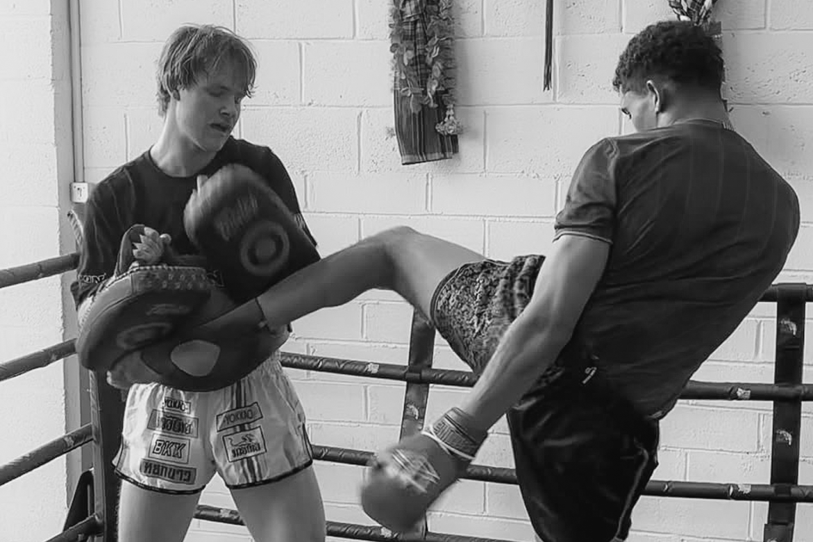 Two people training in a boxing gym: one holding focus mitts, the other performing a kick.