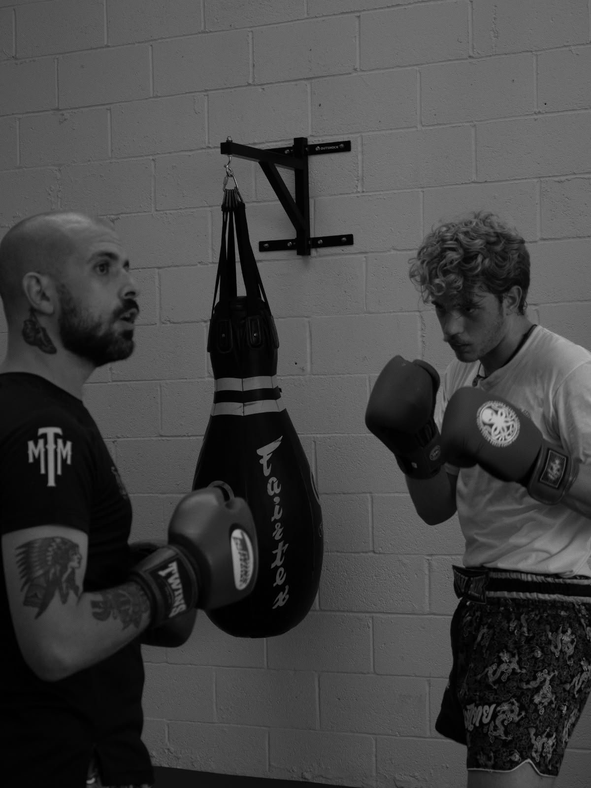 Two men with boxing gloves practicing in a gym, standing near a against a brick wall with a punching bag hanging from the ceiling.