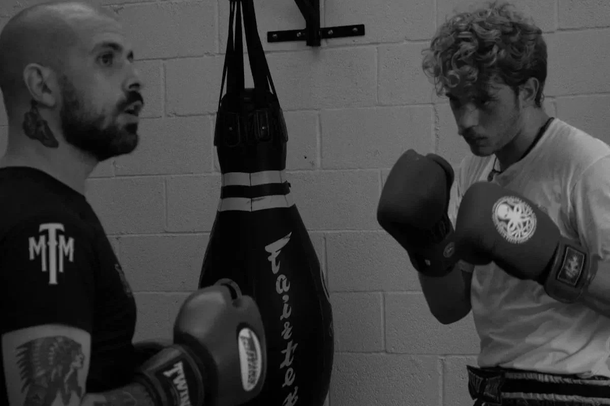 Two men in a boxing gym, one with a beard and tattoos, wearing boxing gloves, and the other with curly hair and boxing gloves, standing near a punching bag.