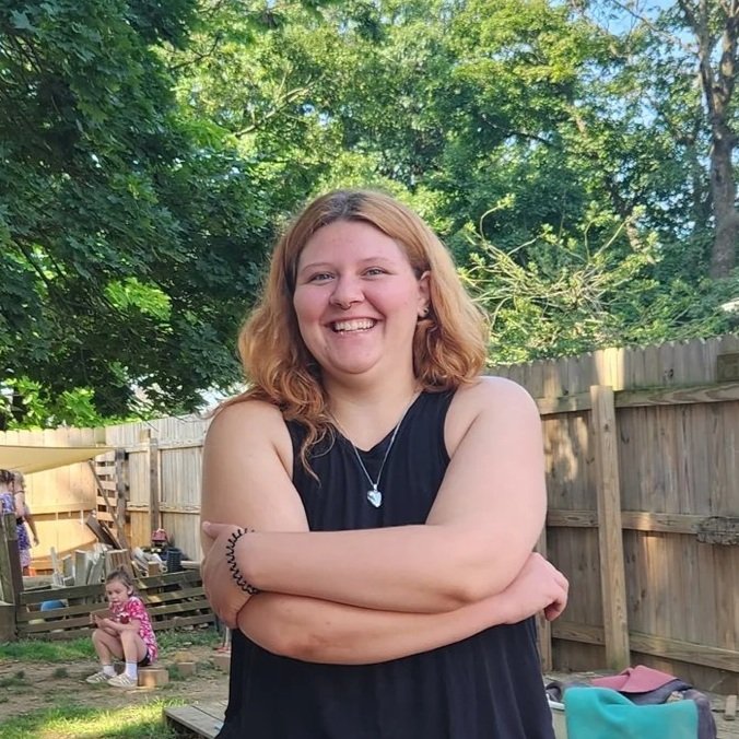 A young woman with shoulder-length red hair smiling and crossing her arms, standing outdoors in a backyard with trees and a wooden fence. Two children are sitting on a small log in the background.