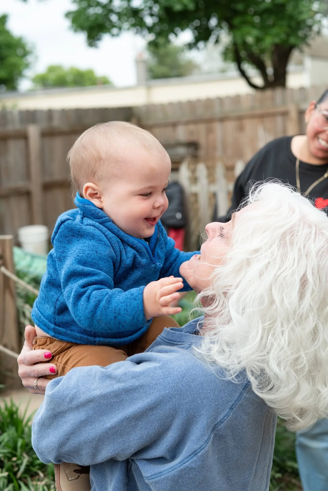 A young boy in a blue hoodie smiling and holding an elderly woman with white hair face to face outdoors, surrounded by a wooden fence, trees, and gardening supplies.