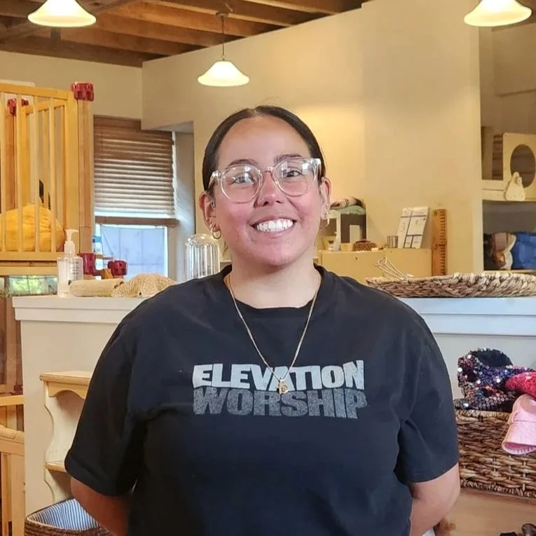 A woman with glasses smiling, wearing a black t-shirt that says 'ELEVATION WORSHIP,' standing inside a cozy room with wooden ceiling beams, shelves, and a window with blinds.