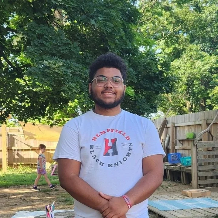 Young man with glasses and beard smiling outdoors in a backyard with trees, wooden fence, and a girl in the background.