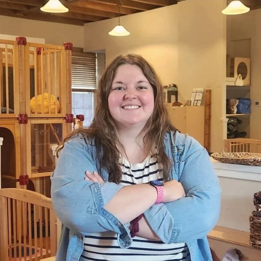A woman with long wavy brown hair smiling with her arms crossed in a cozy room with wooden furniture, hanging lights, and shelves.