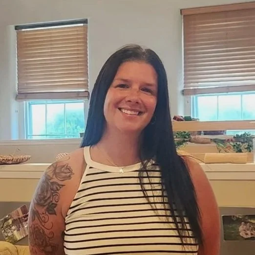 A woman with long dark hair and tattoos on her left arm, smiling in a room with beige walls, wooden blinds, and windows with blue skies and trees outside.