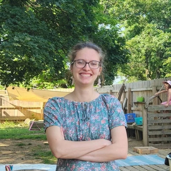 A young woman with glasses, smiling, arms crossed, outdoors in a backyard with trees and a wooden fence in the background.
