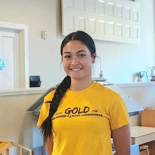 Young girl with long black hair in a braid wearing a yellow T-shirt, standing indoors in front of a counter.
