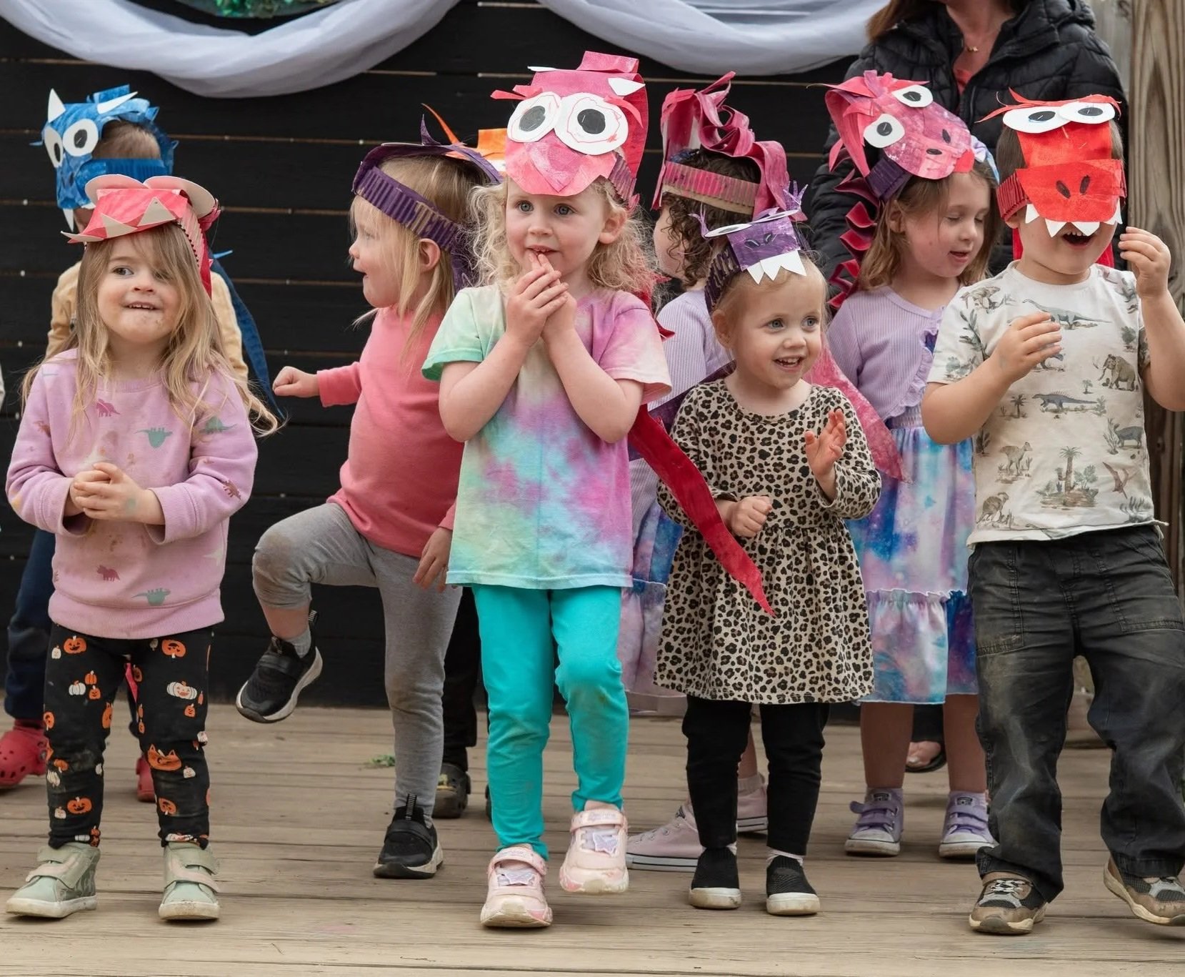 A group of children wearing colorful animal-themed paper masks and casual clothing, standing on a wooden stage, participating in a costume or play activity.