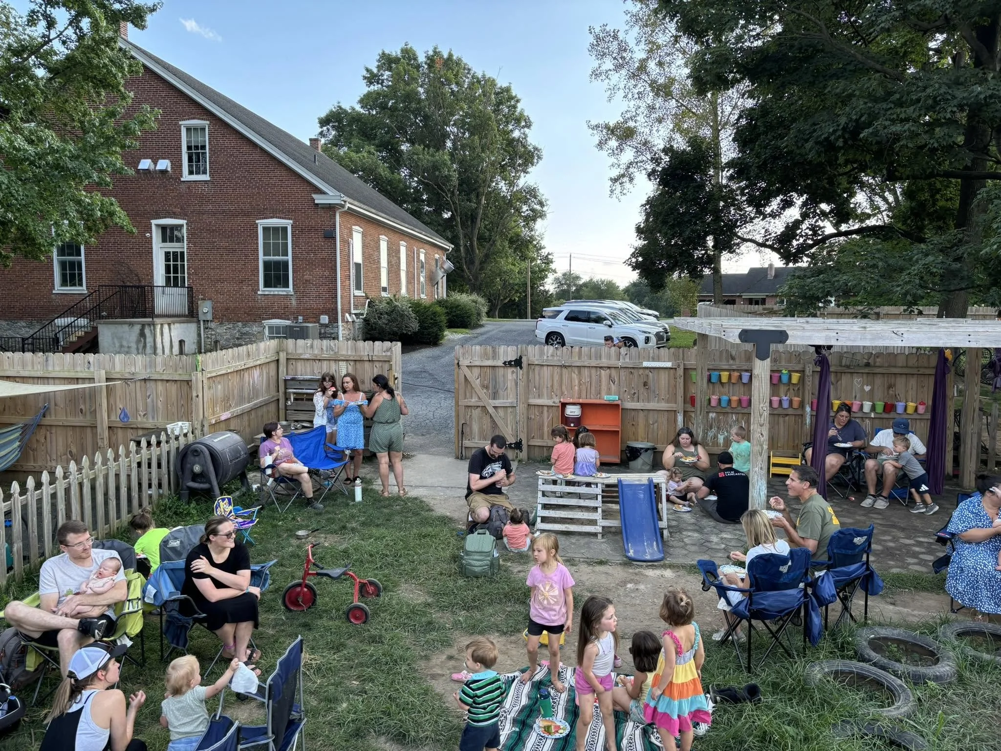 A backyard gathering with children and adults, featuring a wooden fence, a small playground with a slide, and people sitting and standing around. In the background, a large brick house and trees are visible.