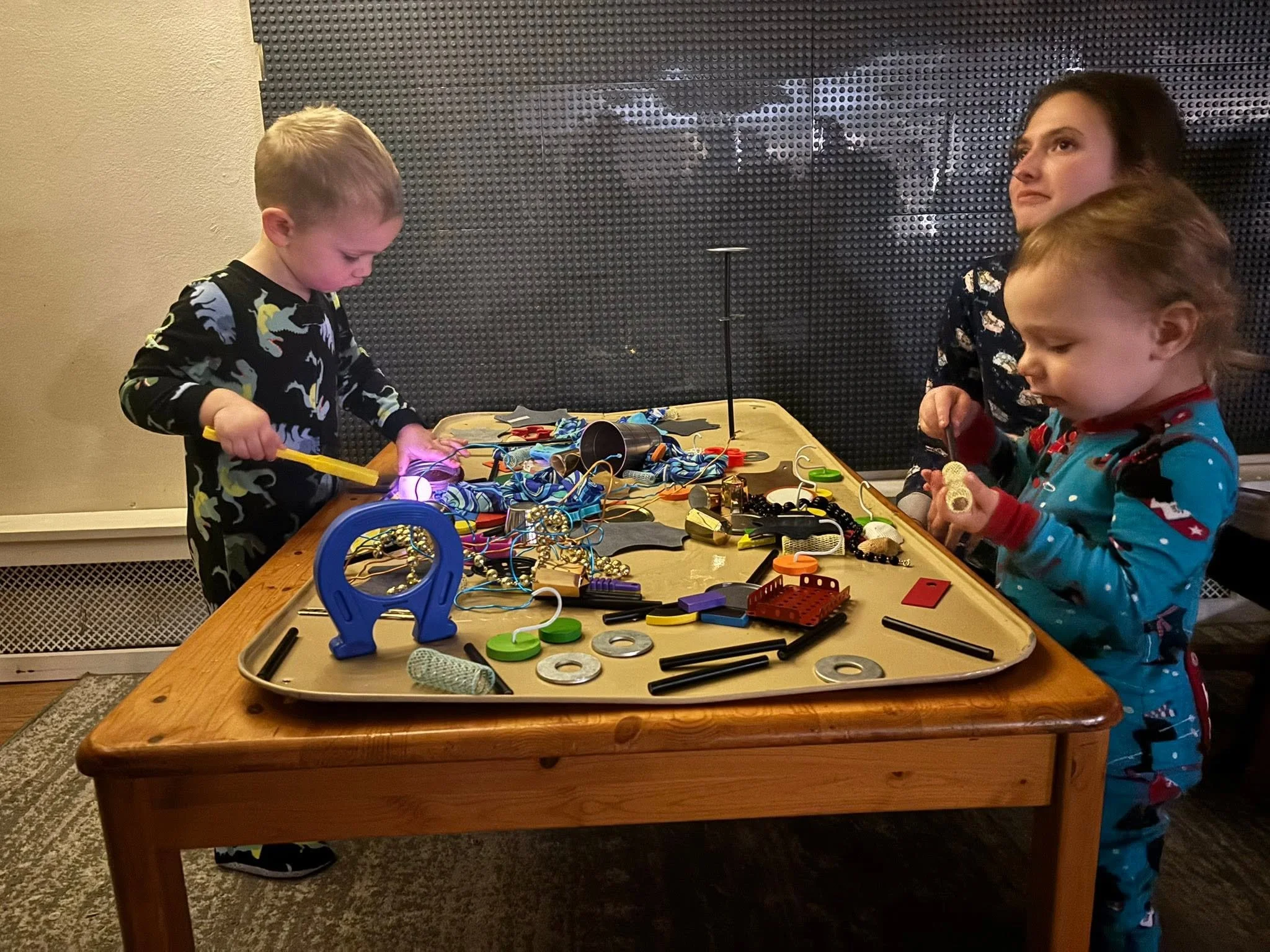 Three children around a table with electronics and craft supplies, working on a project. One boy is looking down at the table, and two girls are focusing on their work.