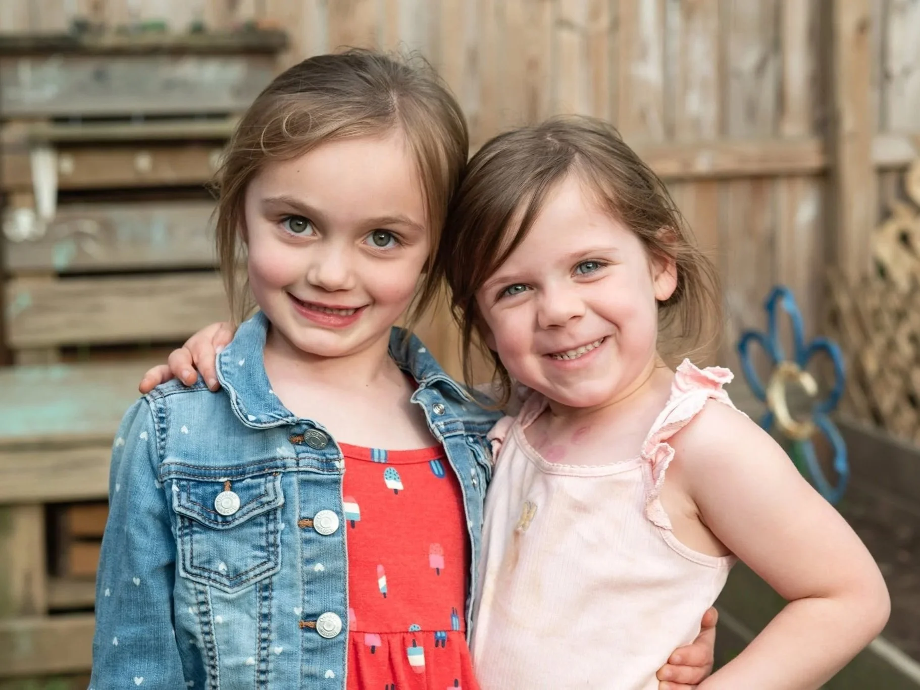 Two young girls standing close together outside, smiling at the camera, with a wooden fence and some children's play equipment in the background.
