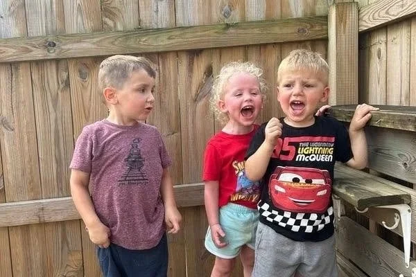 Three young children standing outdoors in front of a wooden fence, two of them are crying and one looks concerned or surprised.