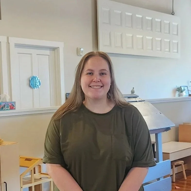 A young woman with long blonde hair wearing a dark green t-shirt, smiling and standing indoors.