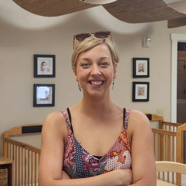 A smiling woman with short blonde hair, wearing a colorful sleeveless top and sunglasses on her head, standing indoors with framed photos on the wall behind her.