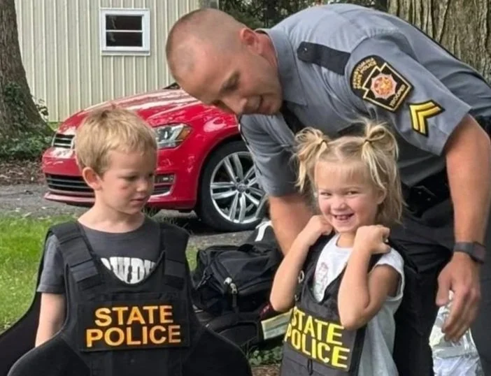 A uniformed police officer smiling and talking with two young children wearing black vests labeled 'State Police.' One child, a girl with blonde hair in pigtails, is smiling and has her fists raised in a playful pose. The boy is looking at the girl with a neutral expression. They are outdoors, with a red car and a building in the background.