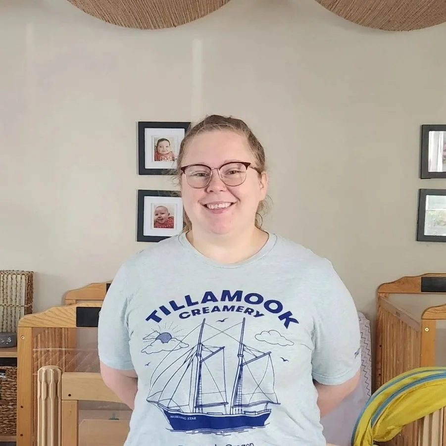 A woman with glasses and a gray T-shirt standing indoors, smiling at the camera. The T-shirt has a ship illustration and reads 'Tillamook Creamery.' In the background, there are framed photos on the wall and wooden furniture.