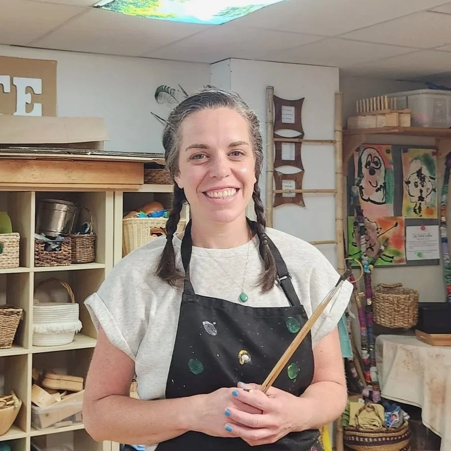 A smiling woman with braided hair and a paint-stained apron holding a paintbrush in an art studio, surrounded by shelves of art supplies and colorful paintings.