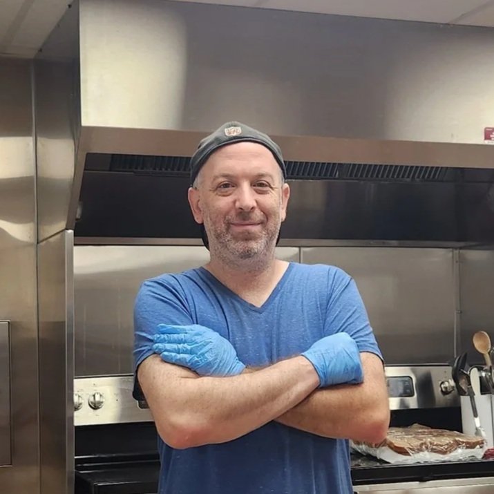 A man wearing a backwards cap, blue shirt, and blue gloves standing in a commercial kitchen in front of a stove, with arms crossed and smiling at the camera.