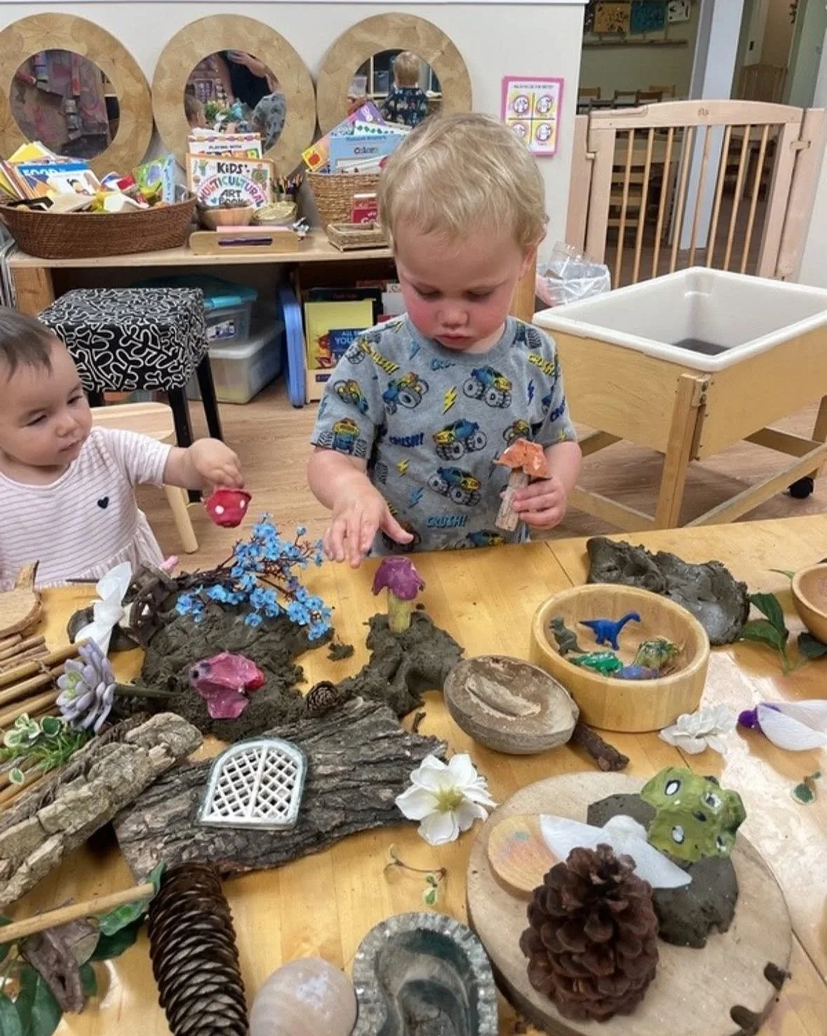 Two young children at a table in a classroom, playing with various natural and craft materials like twigs, pine cones, stones, and small figurines. The room has educational toys and storage units in the background.