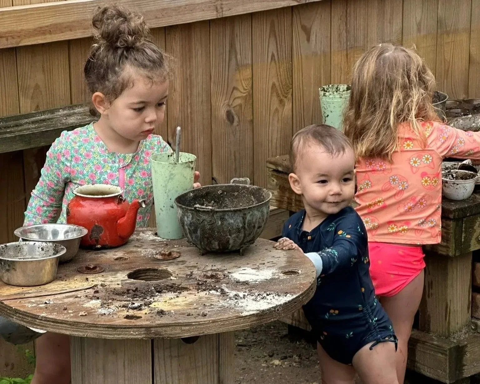 Three young children engaged in a messy outdoor activity at a wooden table, with various bowls and tools, in a backyard with wooden fencing.