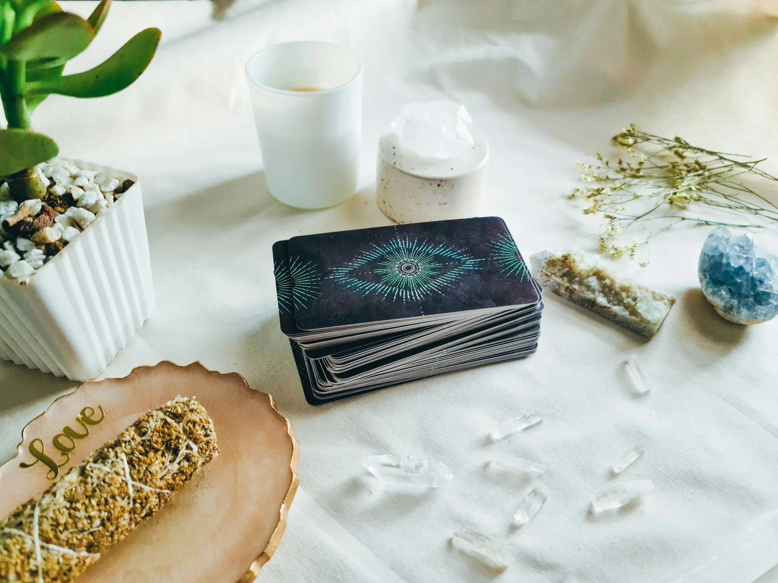 Decorative setup with a potted plant, tarot card deck, a candle in a white holder, a small container with cream, some dried flowers, a blue crystal, and a plate of granola or baked goods on a white surface.