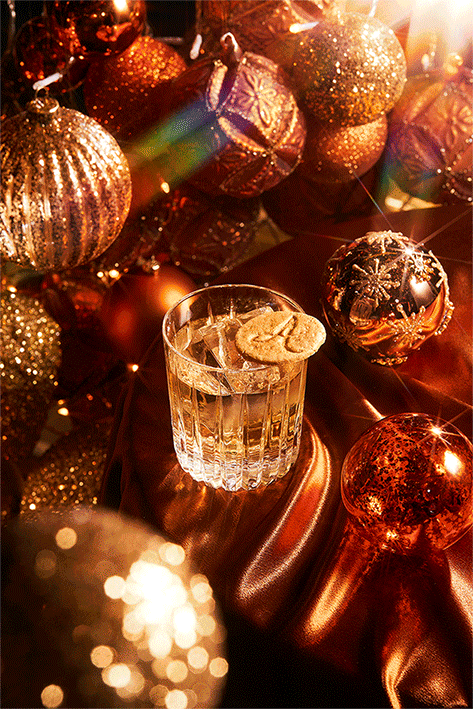 A glass of clear beverage with ice and cookies on top, surrounded by gold and silver Christmas ornaments on a shiny satin surface.