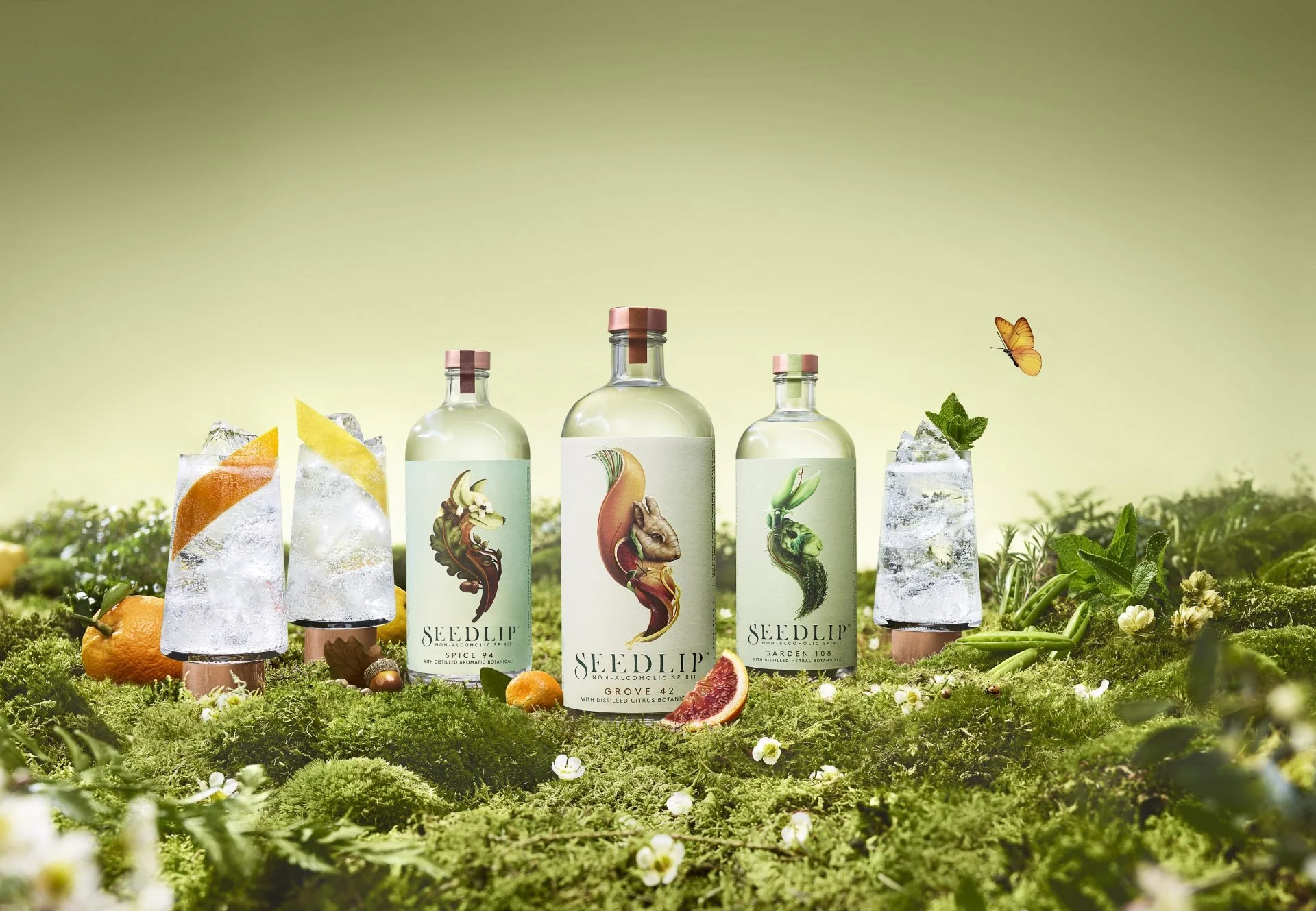 Three bottles of Seedlip non-alcoholic spirit placed on lush green moss with figs, oranges, and fresh herbs, set outdoors with a butterfly flying nearby.