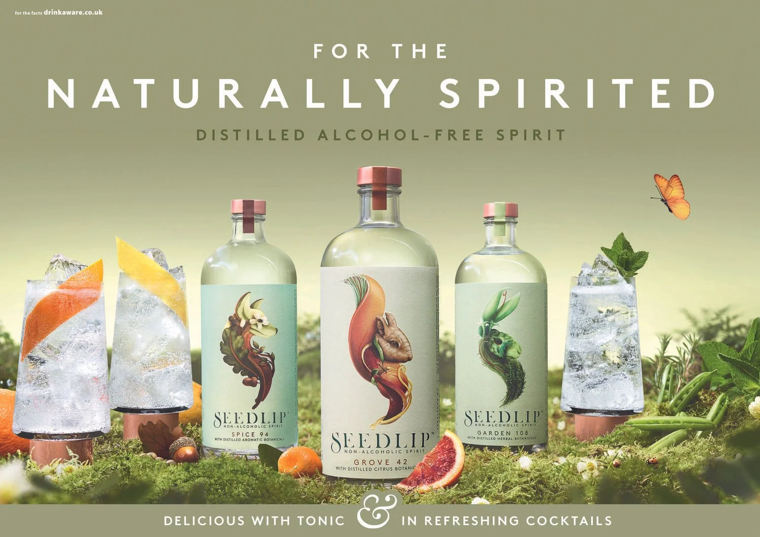 Three bottles of Seedlip non-alcoholic spirit with colorful botanical illustrations on their labels, set among glasses filled with ice and garnished with fruits and herbs, outdoors with greenery and a butterfly in the background.