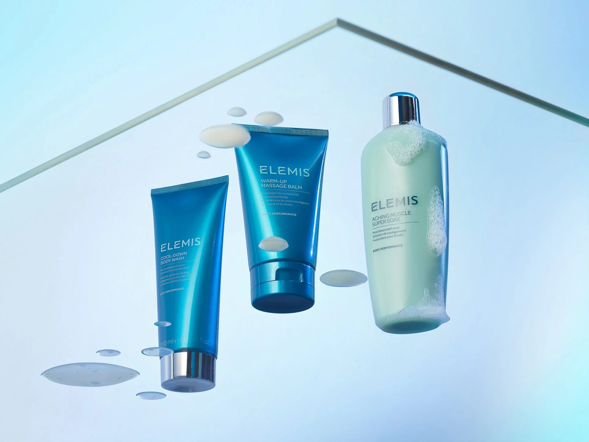 Three blue and green Elemis skincare products, including body wash, massage balm, and bath soak, with drops of liquid and foam on a light blue background.