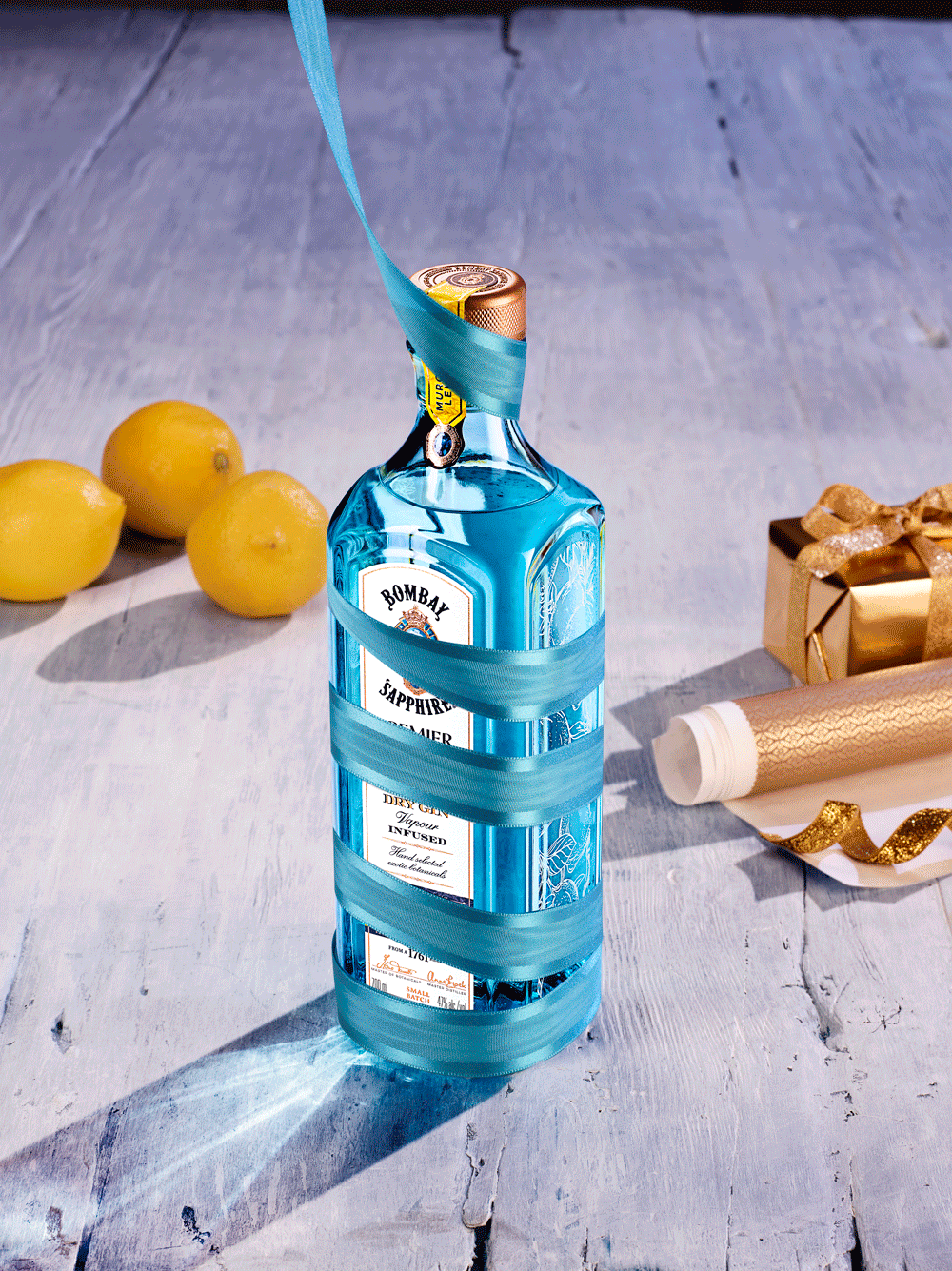 A bottle of Bombay Sapphire gin with a blue ribbon wrapped around it, three lemons to the left, and a small wrapped gift and a gold ribbon on the right, all on a light wooden surface.